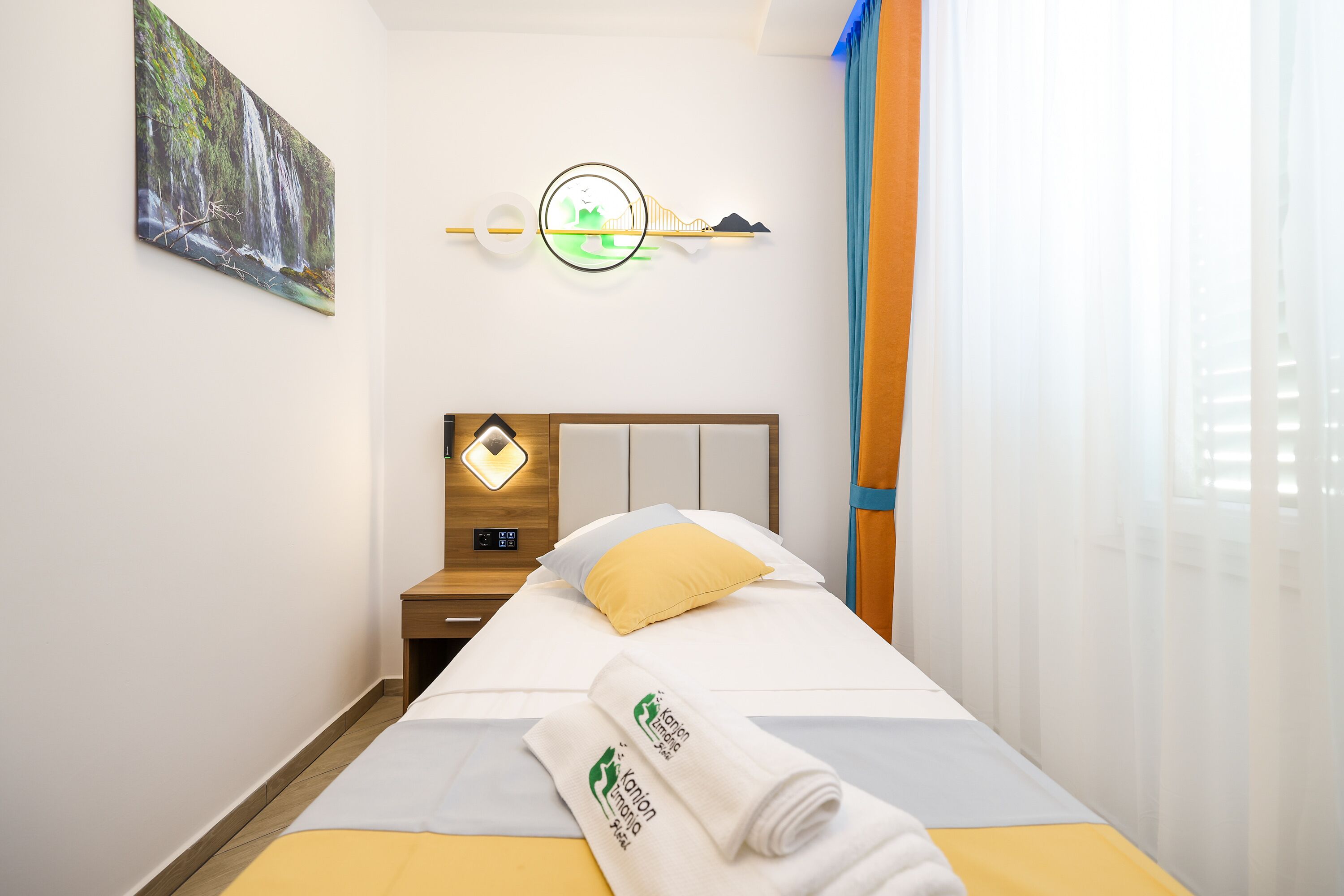 Una habitación de hotel con cama, mesita de noche, un cuadro, una lámpara de pared y una ventana con cortinas.