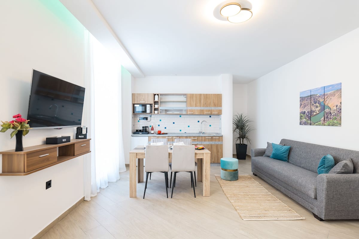 Un salon moderne avec un canapé gris, un téléviseur à écran plat et un coin repas comprenant une table et des chaises en bois.
