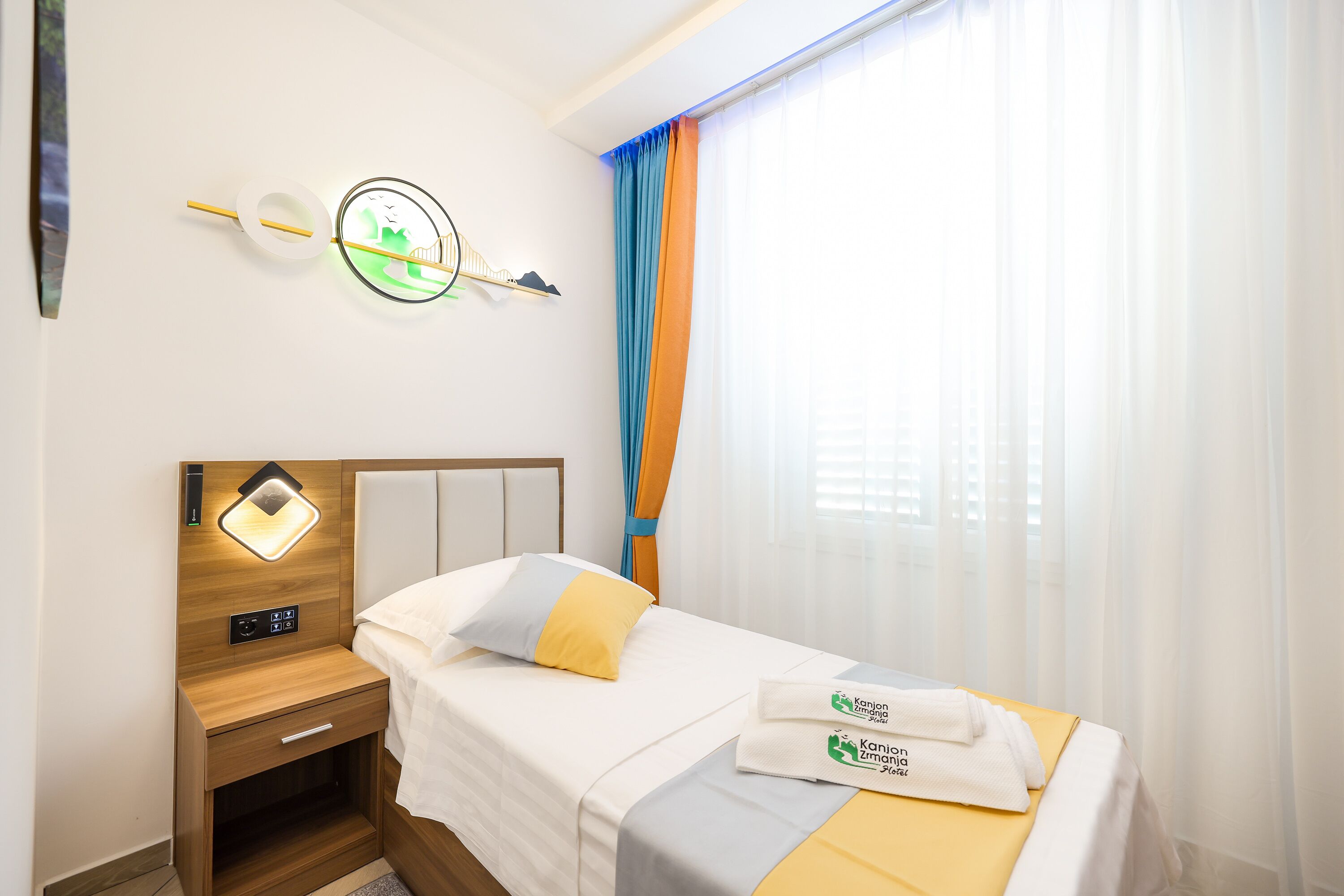 Quarto de hotel com cama, mesa de cabeceira, relógio na parede e janela com cortinas.