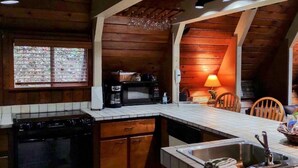 A kitchen with wooden cabinets, a stove, a microwave, and a coffee maker. There is a window with blinds, a lamp, and a chair. - A River Retreat is an Authentic A-Frame with 200' frontage on the Toccoa River (Blue Ridge)