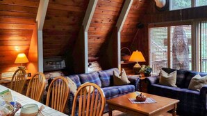 A cozy cabin interior with a high ceiling, wooden beams, a dining area with a table and chairs, and a sofa set. - A River Retreat is an Authentic A-Frame with 200' frontage on the Toccoa River (Blue Ridge)