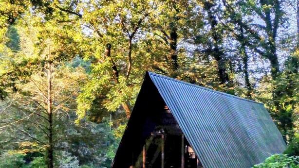 A cabin with a sloped roof, a deck, and a river in the background. - A River Retreat is an Authentic A-Frame with 200' frontage on the Toccoa River (Blue Ridge)