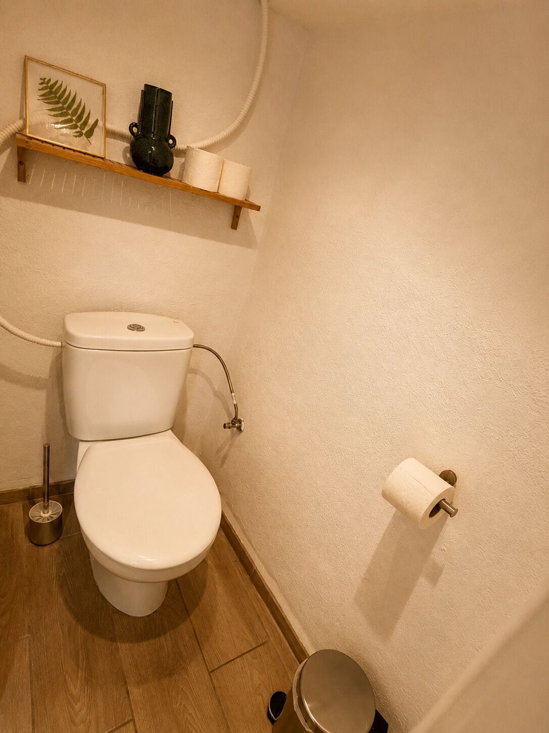 A bathroom with a toilet, a shelf with a framed leaf, a vase, and a toilet paper holder.