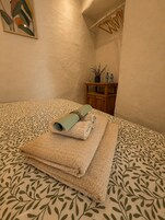 Una cama bien tendida con toallas enrolladas y una pequeña mesita de madera con una planta en maceta y botellas.