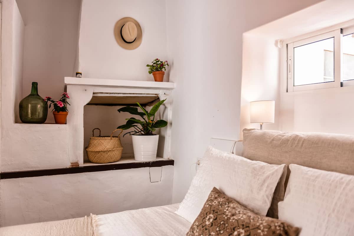 A bedroom with a bed, a shelf with plants, a hat on the wall, and a window.