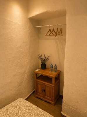 A corner room with a wooden cabinet, a hanging clothes rack, a potted plant, and two water bottles on a small table. - Encanto Guesthouse (Canillas de Aceituno)