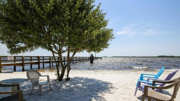 A beach with a wooden dock, a tree providing shade, and beach chairs. - Beach Retreat w/ Pool & Fishing Dock (Port Charlotte)