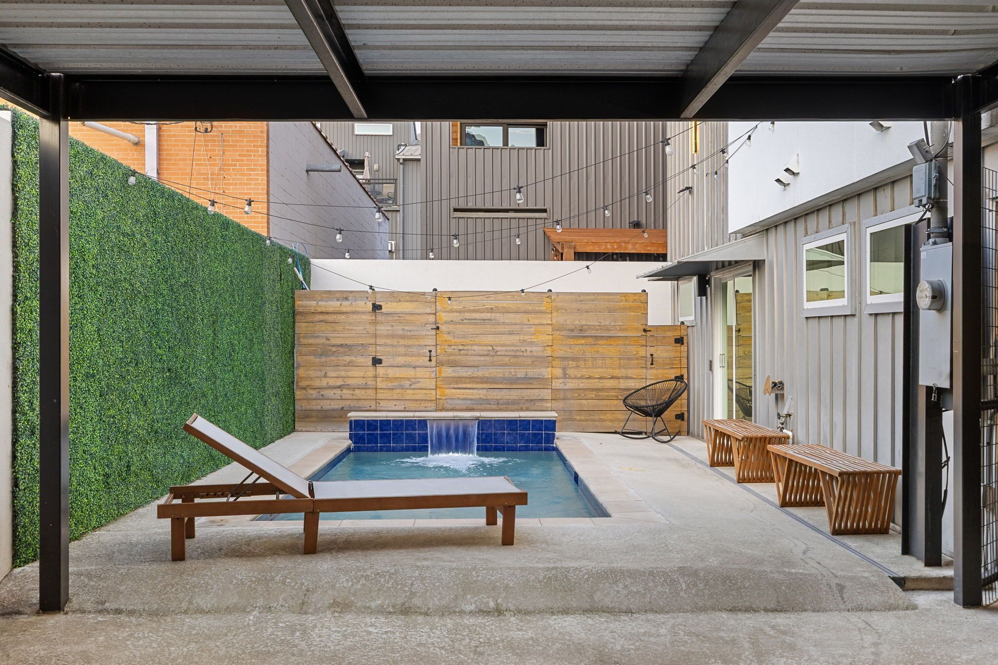 A modern outdoor pool area with a wooden bench and lounge chair, surrounded by a green hedge and a wooden wall.