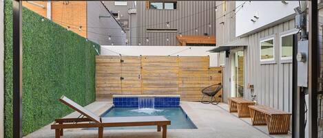 A modern outdoor pool area with a wooden bench and lounge chair, surrounded by a green hedge and a wooden wall.