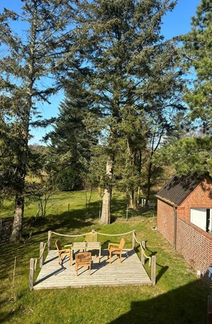 Relax on the deck surrounded by lush greenery and tall trees, with a brick building nearby.