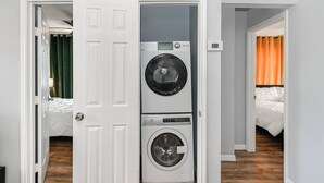A laundry room with stacked washer and dryer units, a door leading to a bedroom with a bed and orange curtains, and a visible thermostat on the wall.