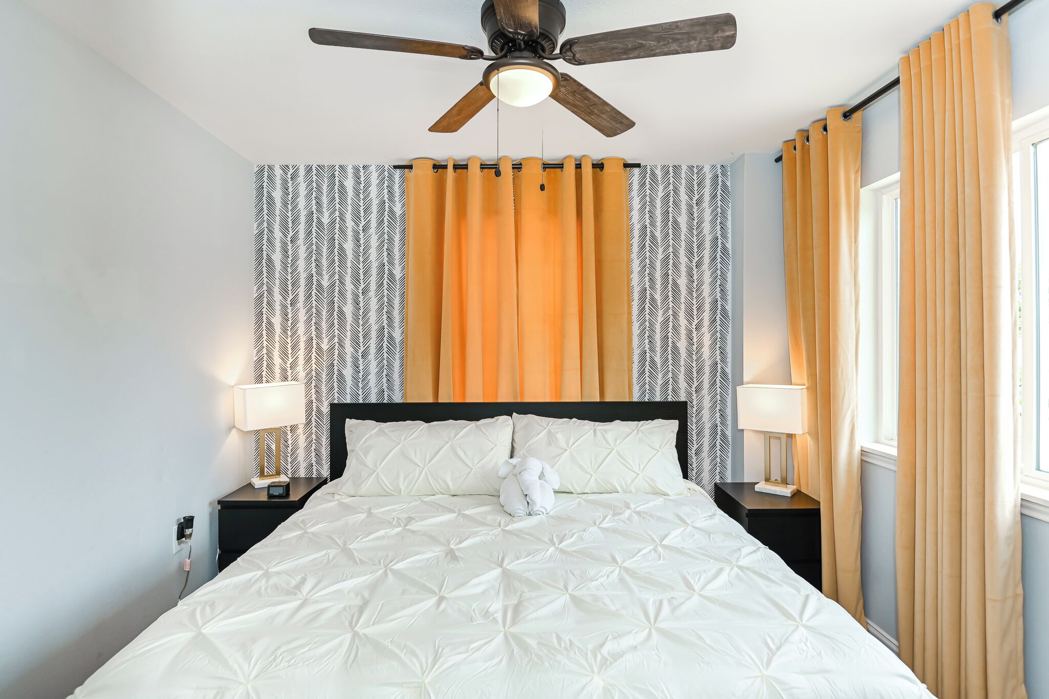 A neatly made bed with a white comforter, a ceiling fan, and a window with curtains.
