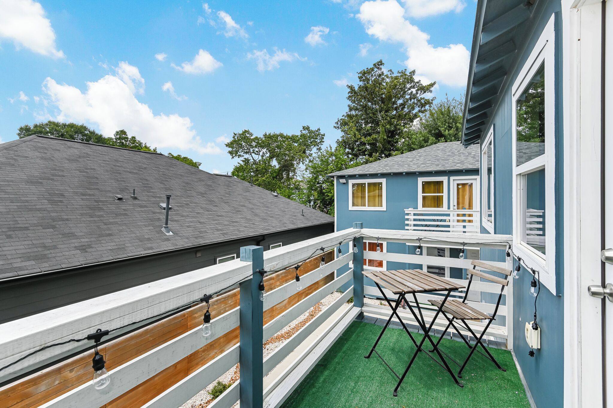 Une maison bleue avec un balcon, une clôture en bois et un espace recouvert de gazon synthétique vert, avec une table et des chaises.