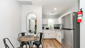 A compact kitchen with white cabinets, a stainless steel refrigerator, a microwave, and a sink. There is a round wooden table with four chairs and a large mirror on the wall.