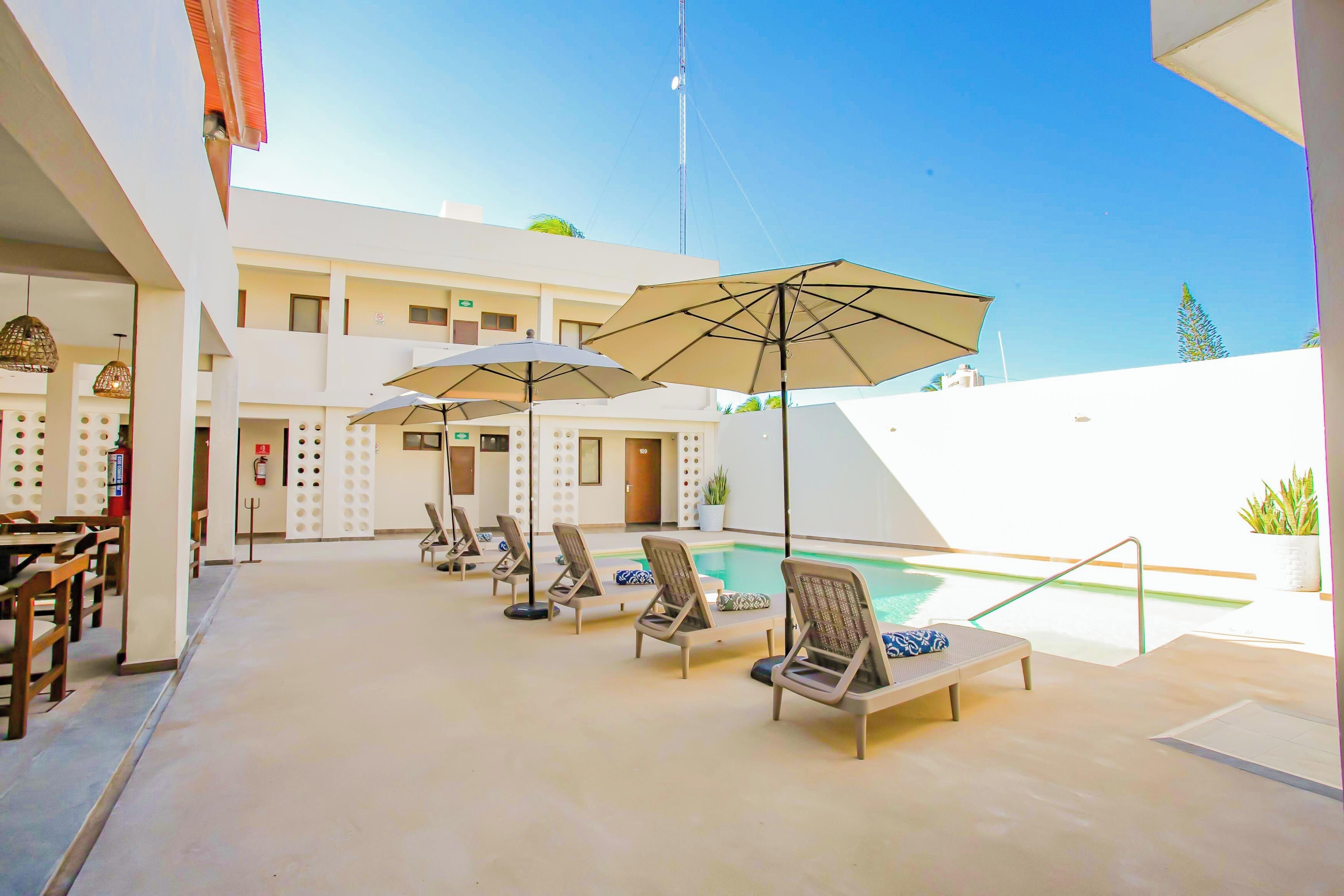 Un espace aménagé au bord de la piscine, avec des chaises longues et des parasols, une piscine, et un bâtiment à plusieurs étages doté de balcons.
