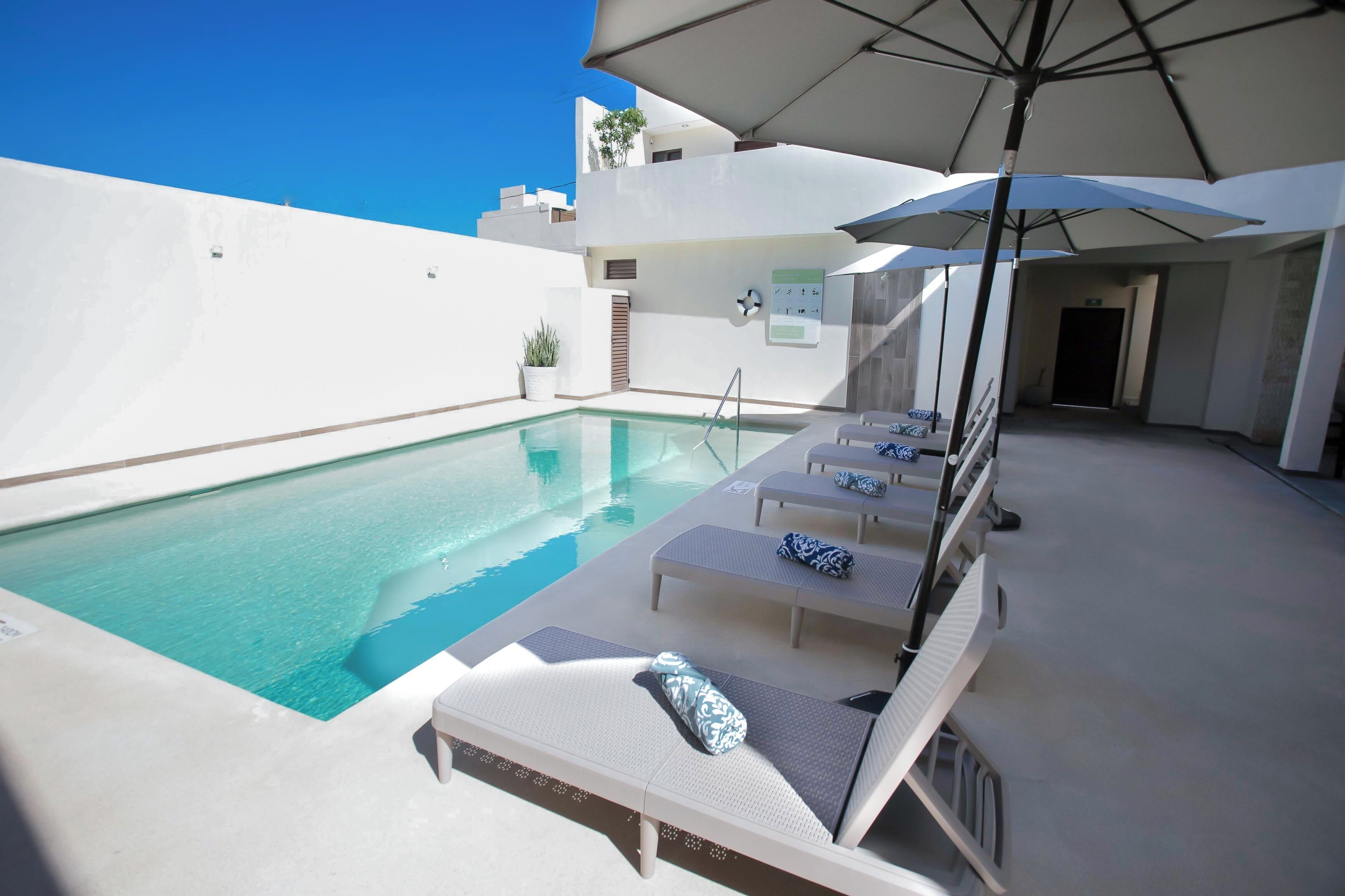 A poolside area with lounge chairs and umbrellas, a clear pool, and a white building in the background.