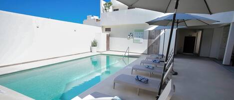 A poolside area with lounge chairs and umbrellas, a clear pool, and a white building in the background.