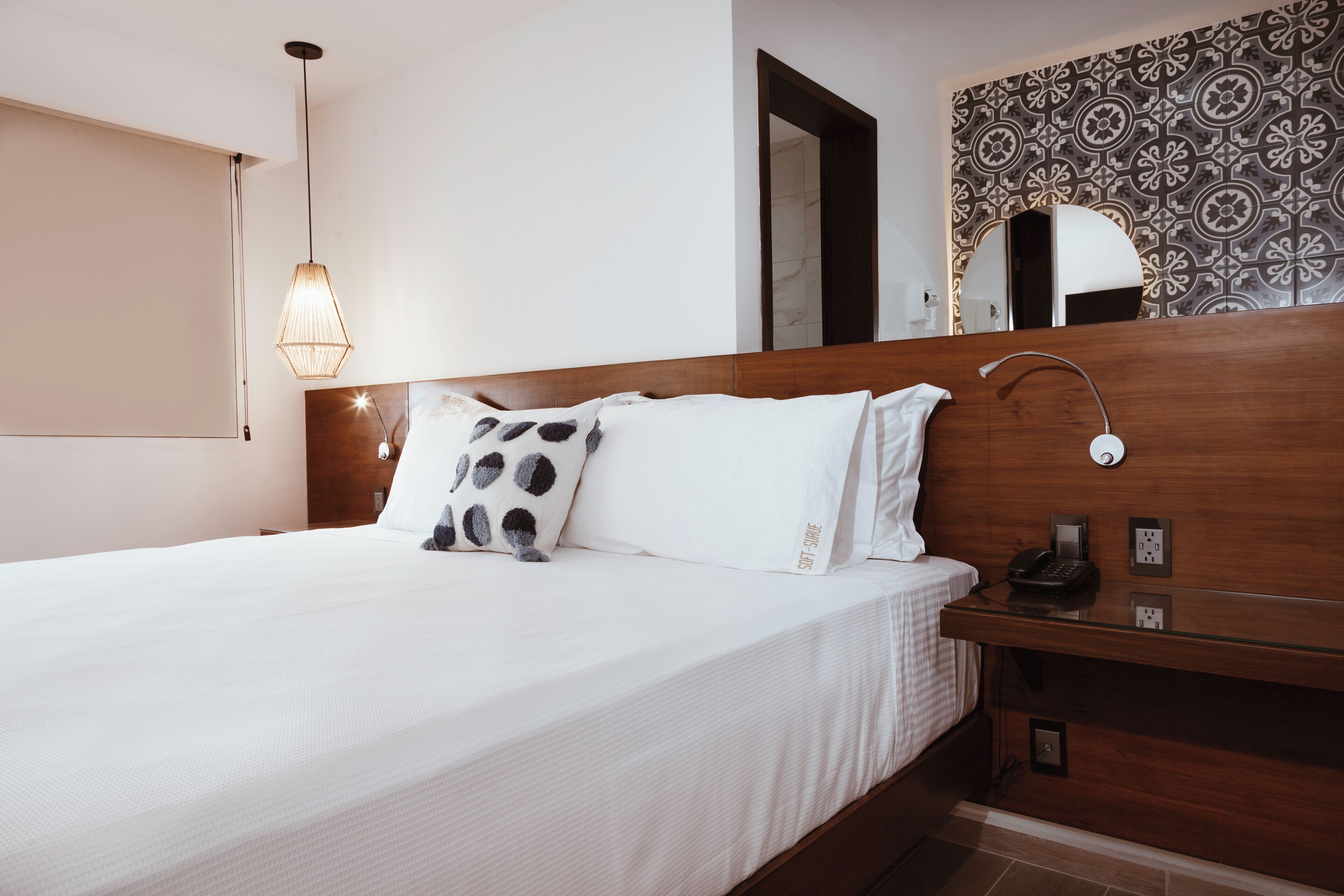 A neatly made bed with white linens and a single black and white pillow. A bedside table with a phone and a lamp is visible. The room features a wooden headboard and a patterned wall.