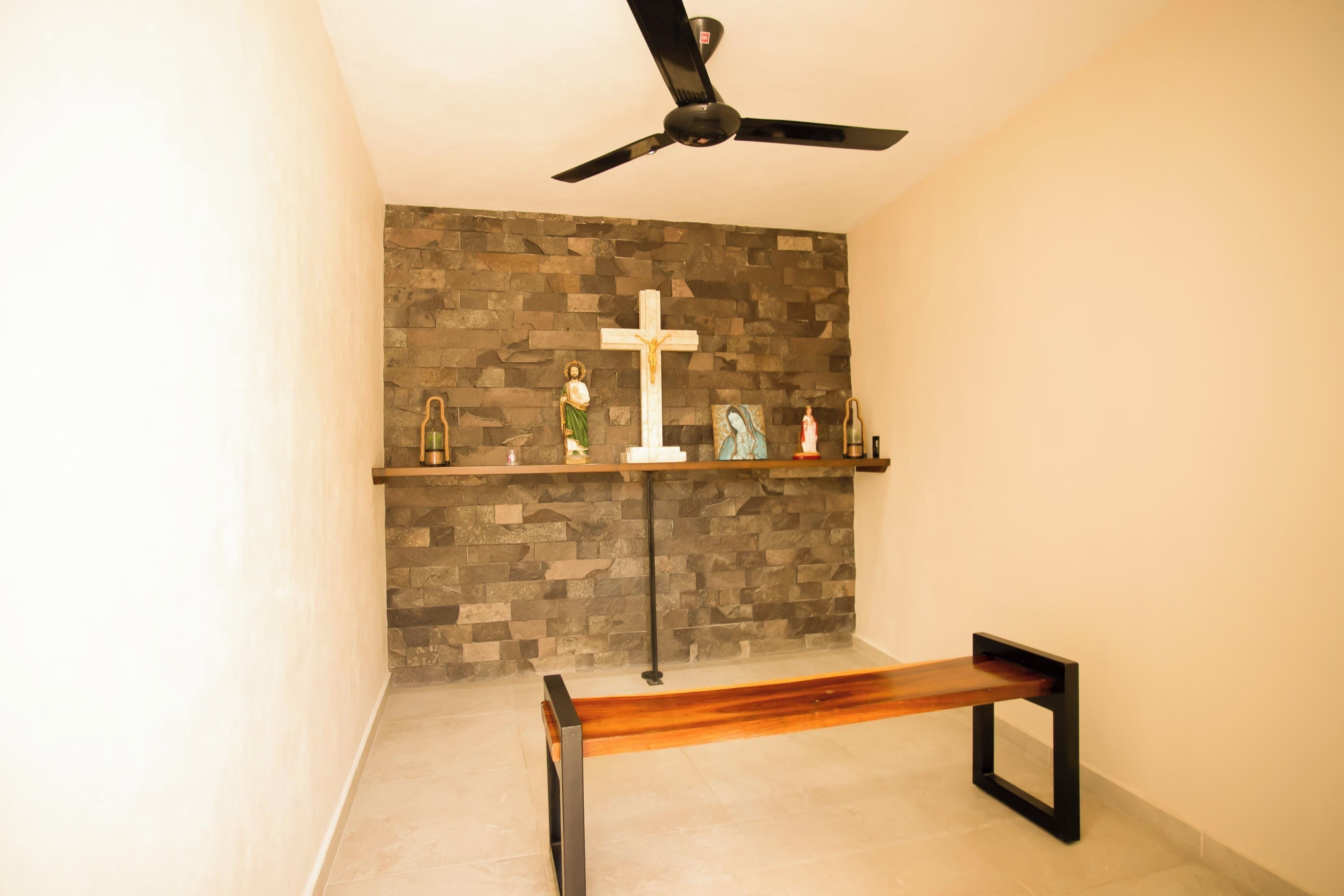 A chapel with a stone wall, a wooden bench, a ceiling fan, and religious statues.
