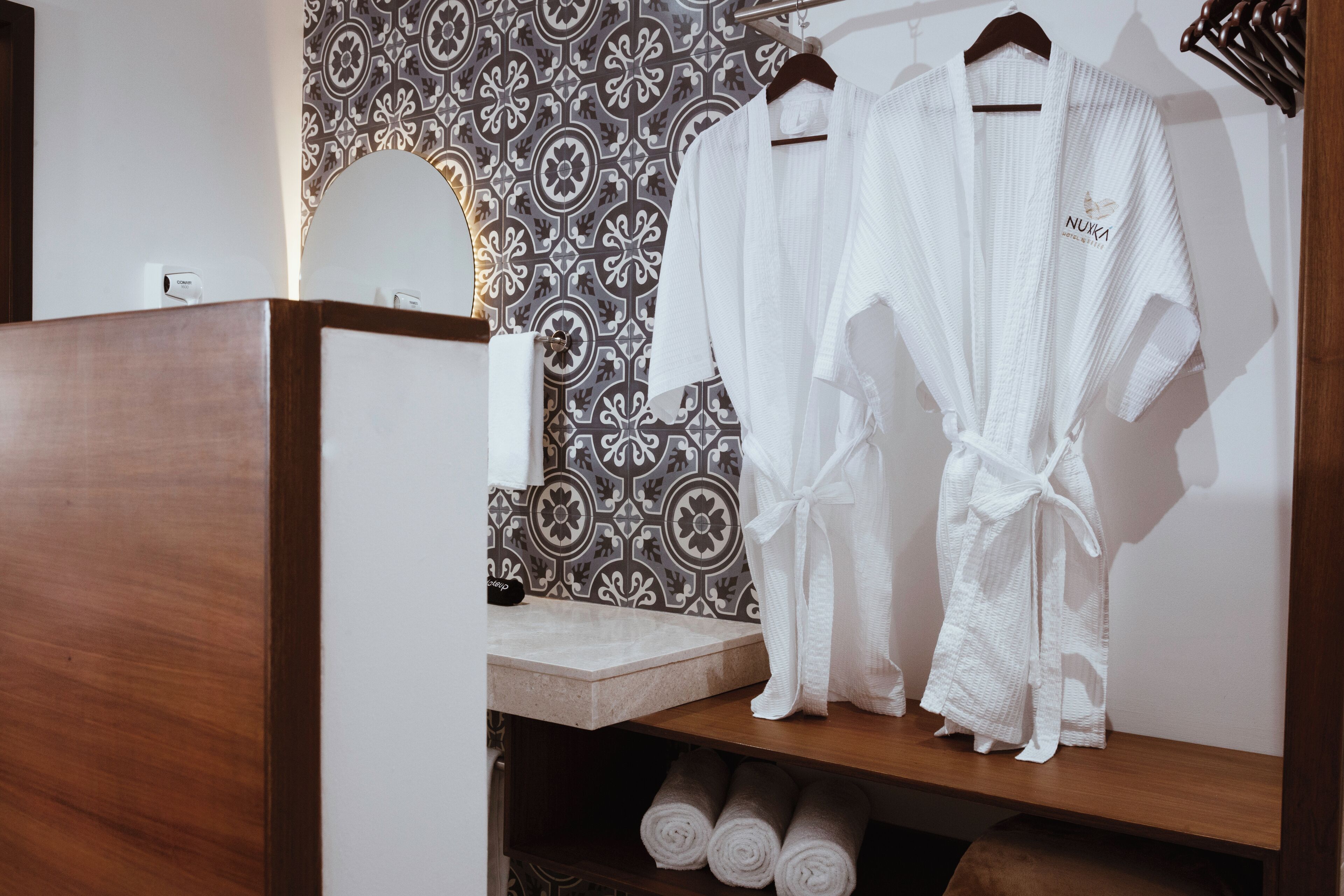 Een badkamer met een houten wastafelmeubel, een behang met patroon en twee witte jassen aan een rek.