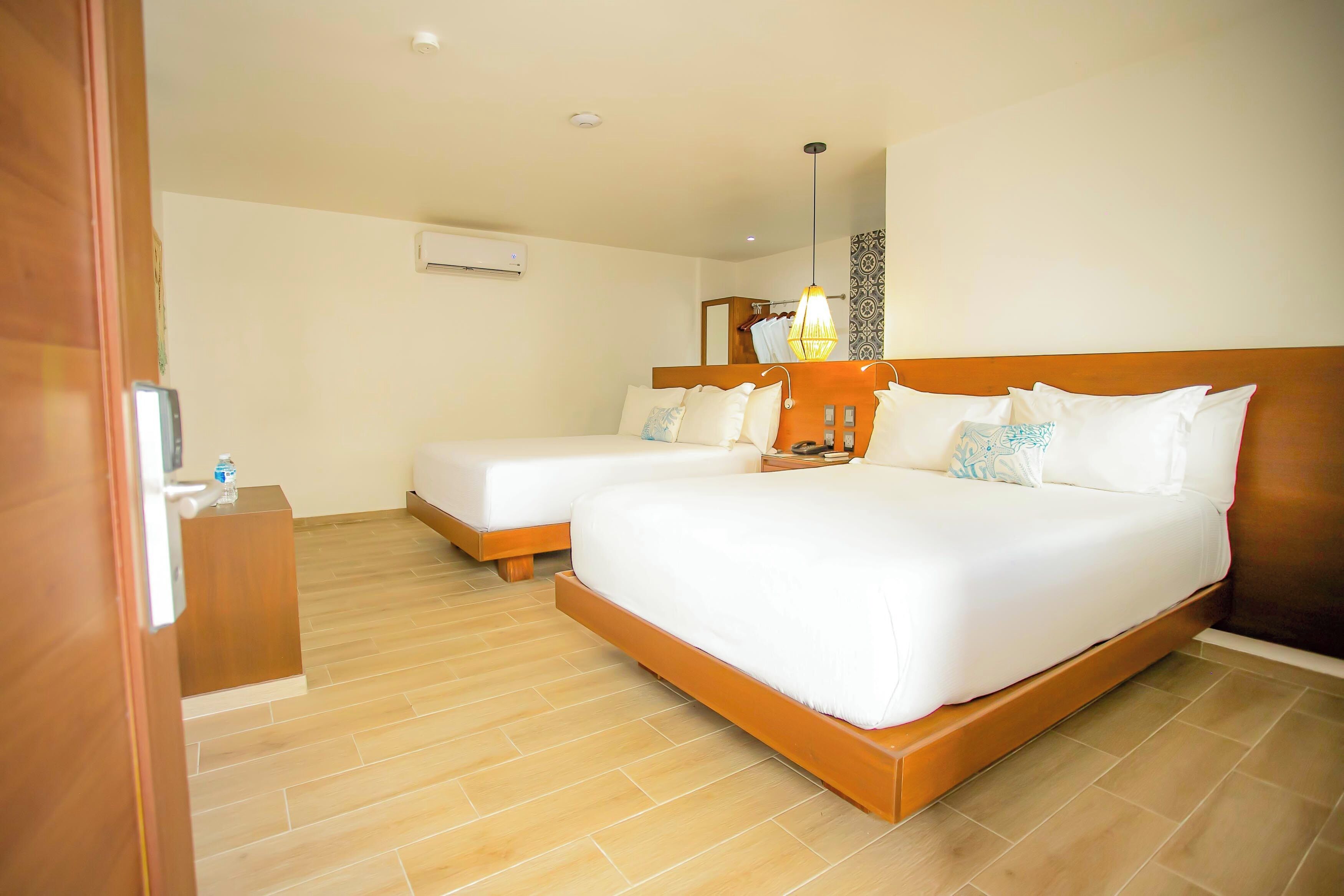 Habitación de hotel con dos camas, cabecera de madera, mesita de noche con lámpara y aire acondicionado instalado en la pared.