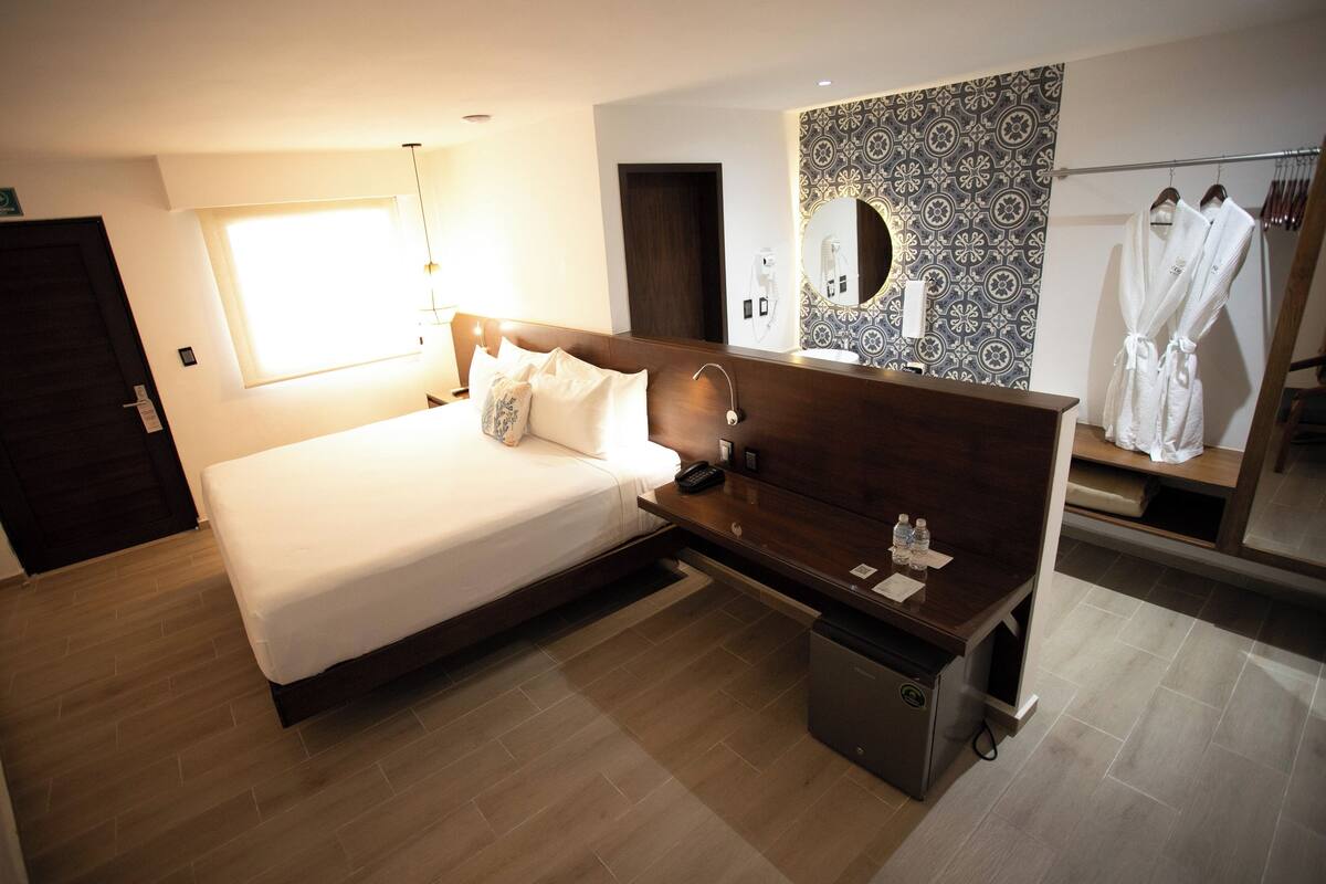 A modern hotel room with a large bed, a bedside table with a lamp, a mirror, and a bathroom with a towel rack.