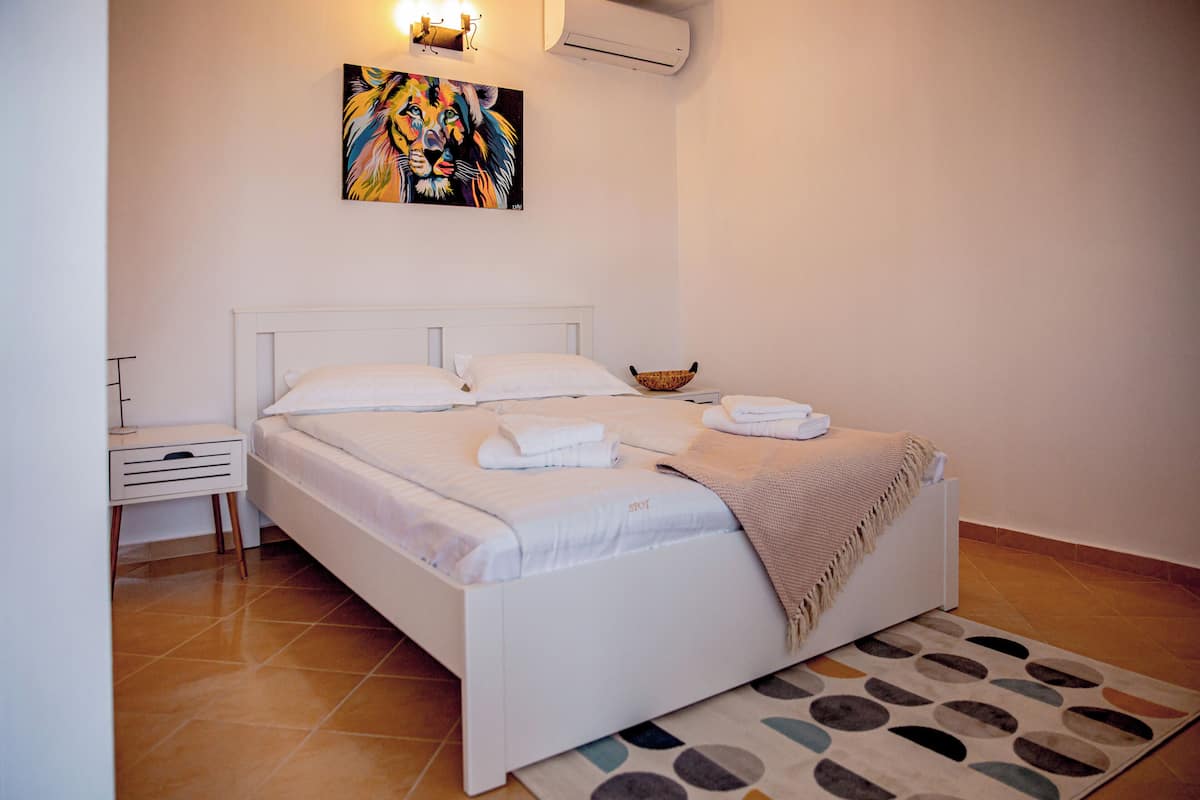 A neatly made bed with white linens and a throw blanket, a bedside table with a lamp, and a colorful abstract painting on the wall.