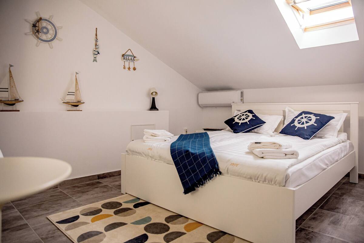 A bedroom with a white bed, nautical-themed pillows, a sailboat decoration, a "RELAX" sign, and a skylight.