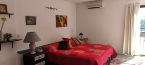 A bedroom with a red quilted bedspread, a bedside lamp, a wall-mounted air conditioner, and a window with curtains.