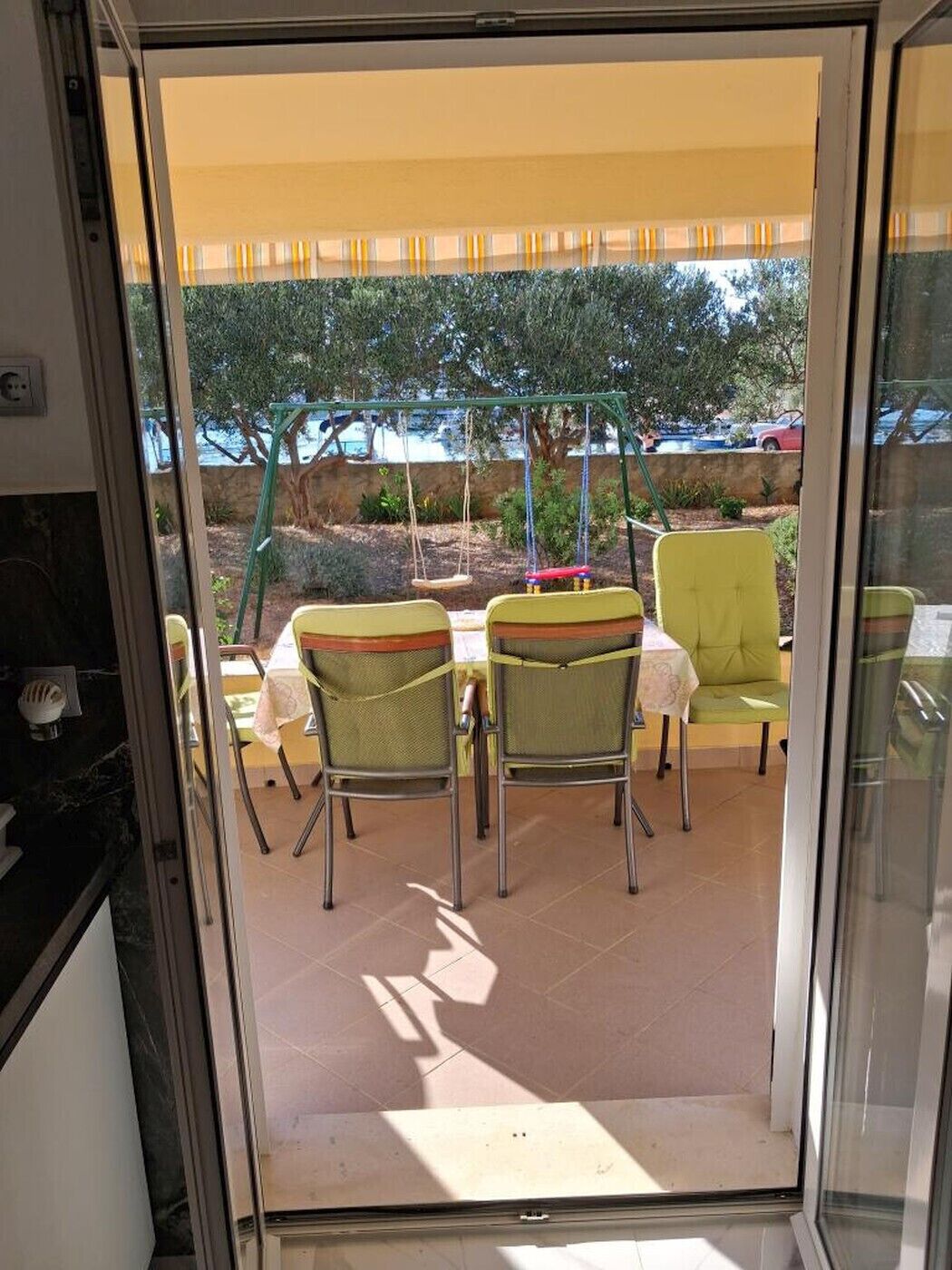 A glass door leading to an outdoor patio with a table and chairs, a swing set, and a view of the sea.