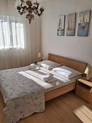 Un lit bien fait, avec des draps blancs et une couverture à motifs floraux, une table de chevet en bois avec une lampe, et trois tableaux représentant des voiliers accrochés au mur.