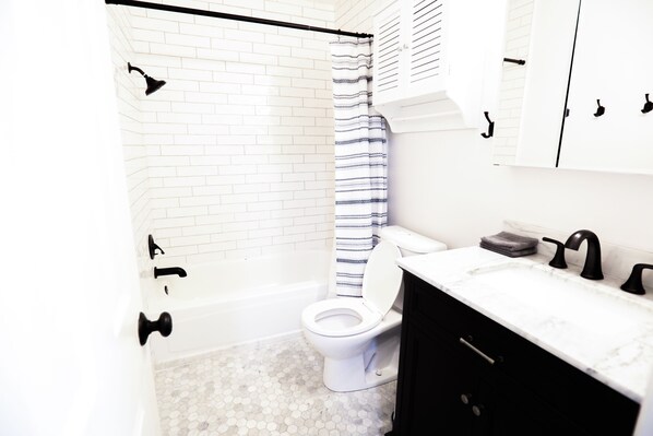 A bathroom with a white toilet, a shower with a striped curtain, a white sink with black faucets, and a tiled floor.