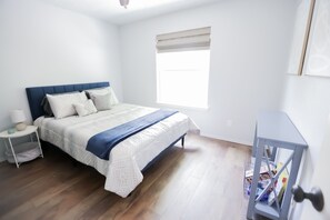 Ein Schlafzimmer mit Bett, Nachttisch, Fenster mit Jalousien und Tür mit kleiner Ablage.