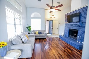A living room with a grey sofa, wooden flooring, a blue brick fireplace, and a ceiling fan.