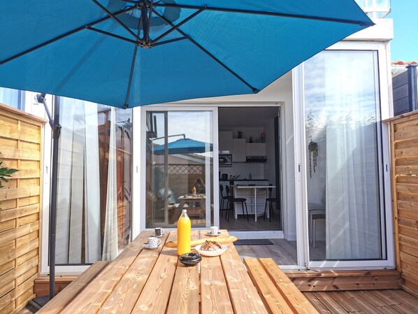 A wooden deck with a table, a blue umbrella, and a glass door leading to a kitchen. - Le P'tit Chaumois 300m from the rocky coast (Les Sables-d´Olonne)