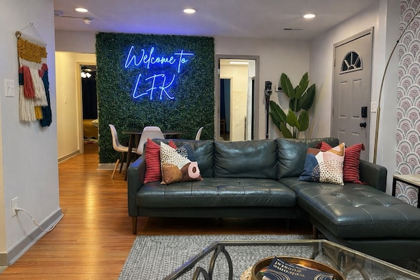 A modern living room with a leather sofa, a neon welcome sign, and a green wall.
