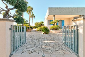 Une allée dallée avec un portail en métal, un bâtiment doté d’une porte bleue et une végétation luxuriante tout autour.