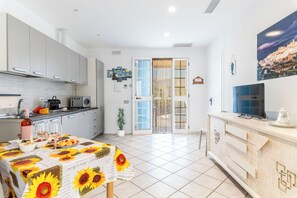 Une cuisine moderne avec des meubles blancs, une table à manger recouverte d’une nappe à motifs de tournesols et un téléviseur posé sur un meuble en bois.