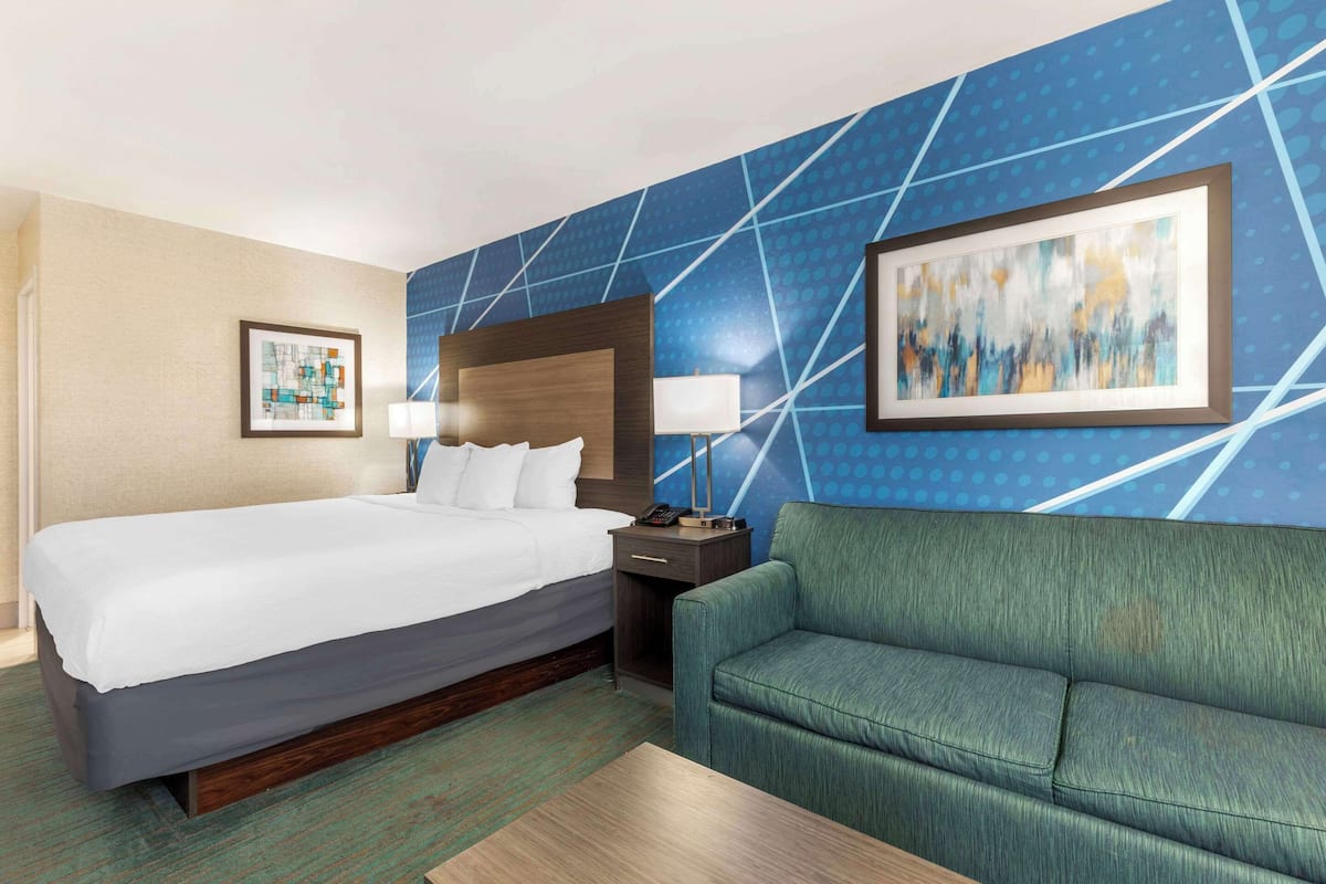 A hotel room with a large bed, a green sofa, a nightstand with a lamp, and two framed artworks on the walls.