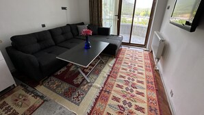 Una sala de estar con un sofá gris oscuro, una mesita con un florero y un tapete estampado, y un ventanal corredizo que da a un balcón.