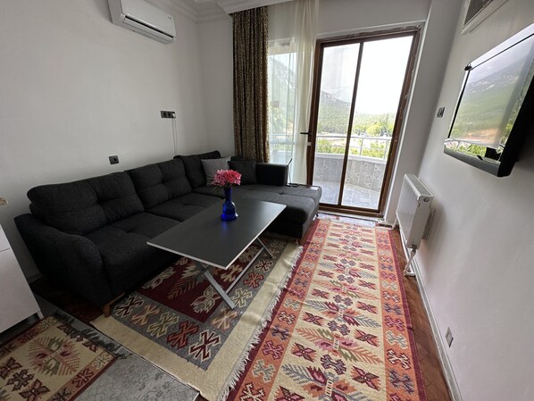 A living room with a dark grey sofa, a small table with a vase of flowers, a patterned rug, and a sliding glass door leading to a balcony. - Olympos Likya Apart Otel (Kumluca)