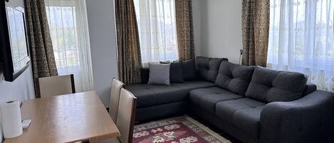 Una sala de estar con un sofá, una mesa de comedor con sillas, cortinas estampadas y un equipo de aire acondicionado.