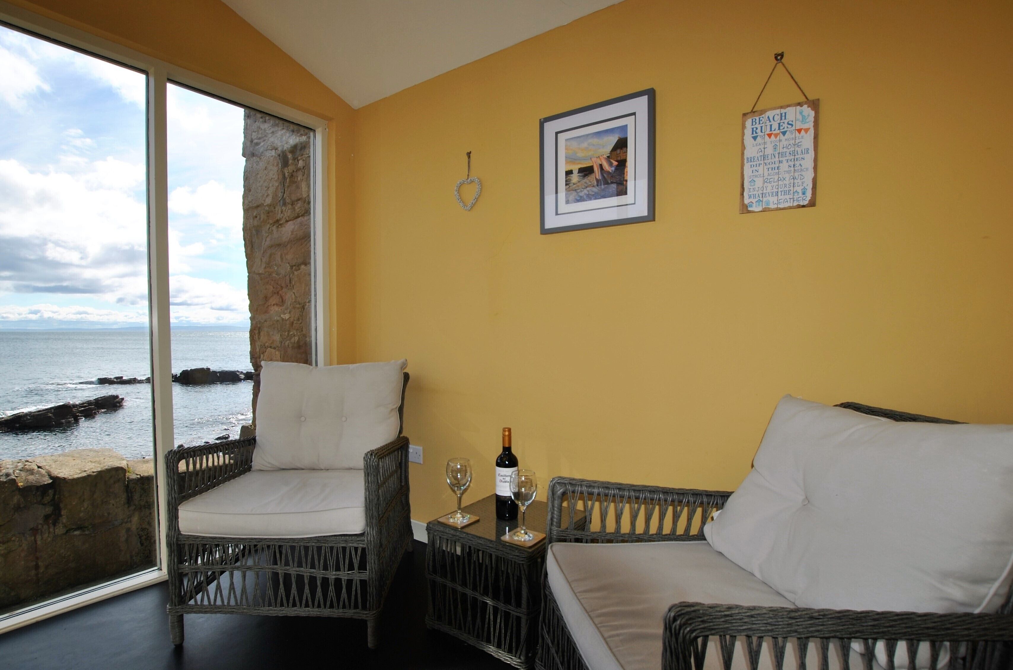 A room with wicker furniture, a glass table with a bottle and glasses, a framed picture, and a sign with beach rules.