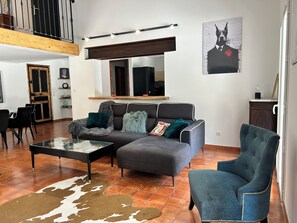 A modern living room with a sectional sofa, a glass coffee table, and a blue armchair. There is a wooden ceiling and a large window. - Propriété de Campagne, Située Entre le Circuit Paul Ricard et Bandol (Le Beausset)