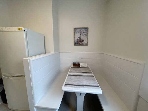 A narrow room with a white bench, a small table, a refrigerator, and a wall-mounted power outlet.