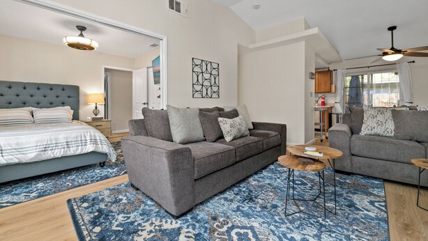 A modern living room with a grey sofa, a blue patterned rug, and a bed with striped bedding. - Casa De Campo (Big Bear City)