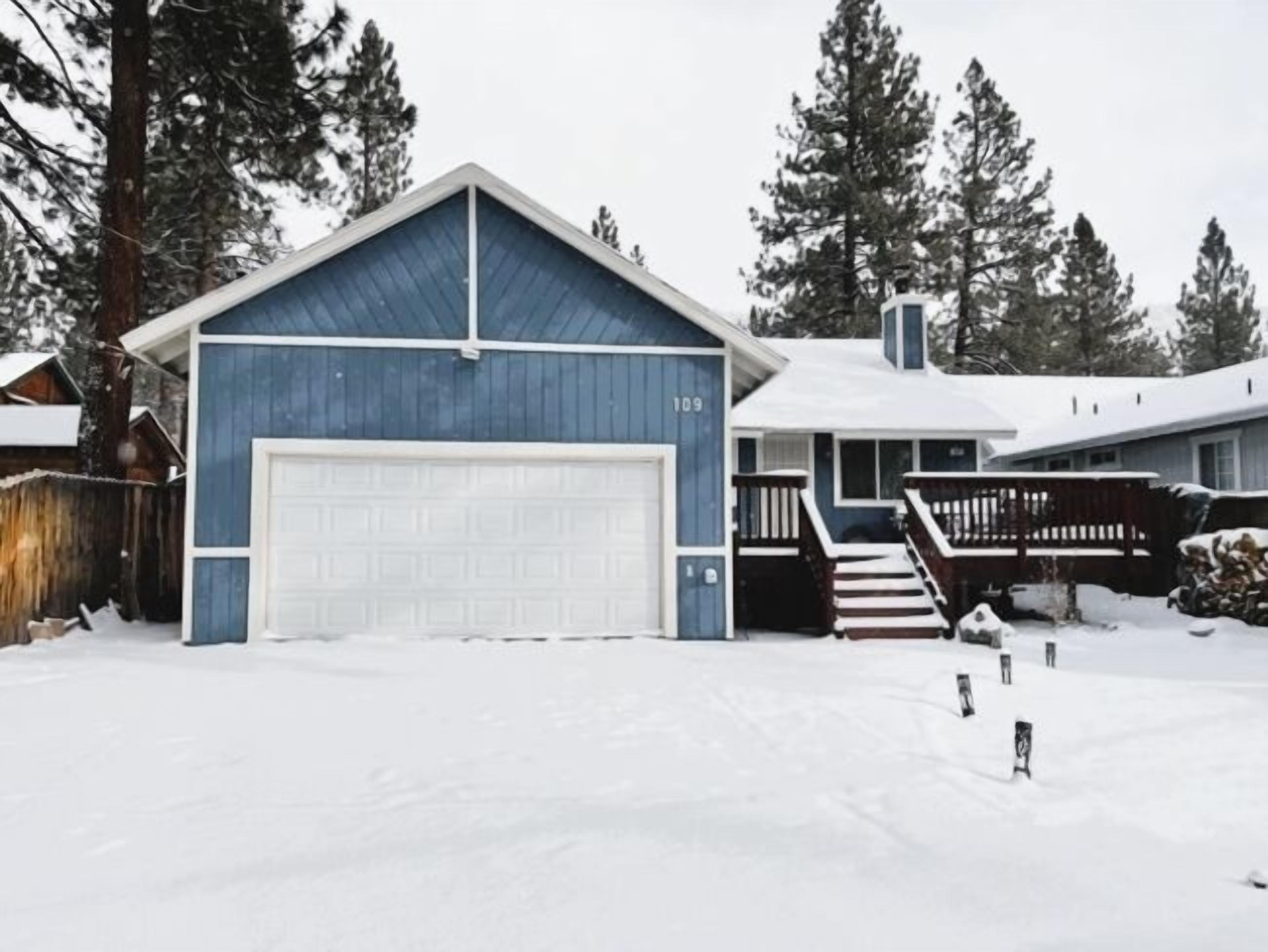 Een huis met twee verdiepingen, met een blauwe garage en een witte voordeur, omgeven door sneeuw en bomen.