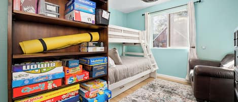 A room with a bunk bed, a bookshelf filled with board games, a brown leather chair, and a large window with curtains.