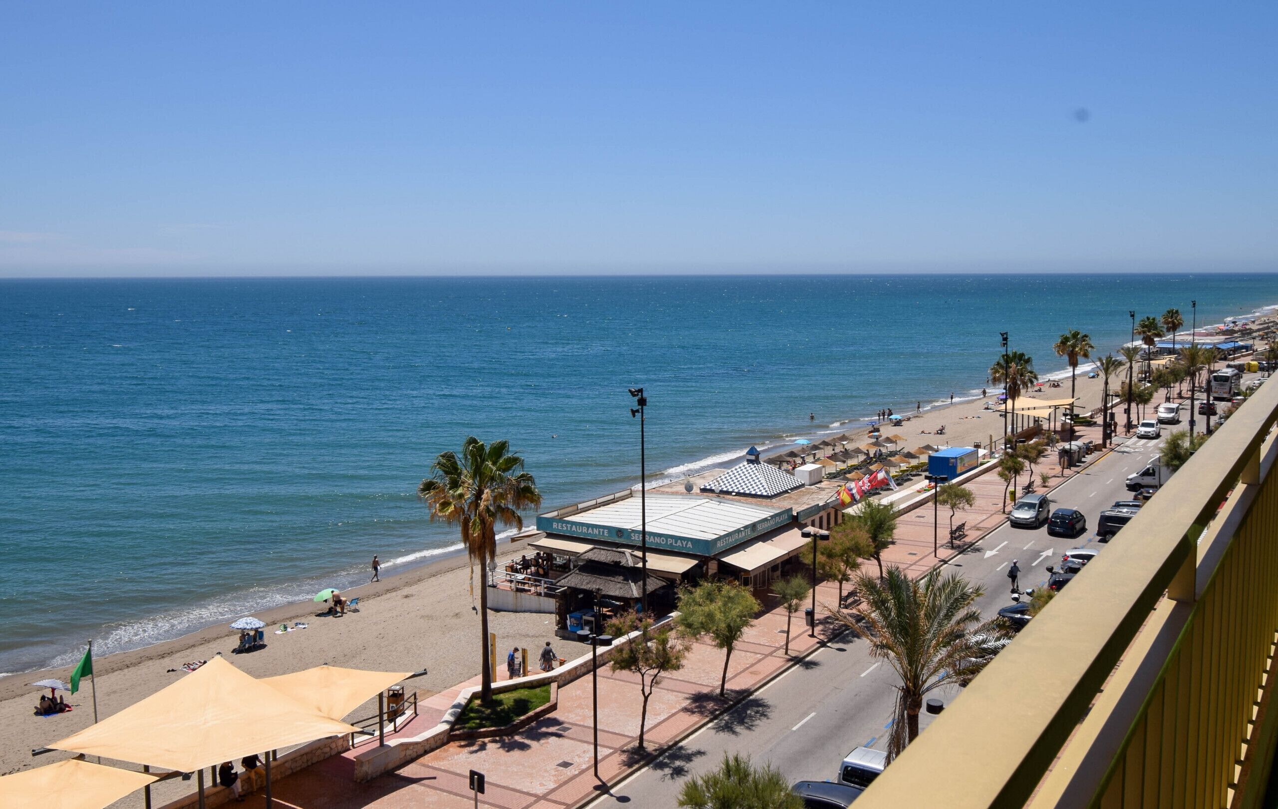 A coastal road with beachside restaurants, palm trees, and a clear view of the ocean.