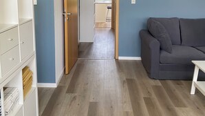 Un salon moderne avec un canapé gris, un meuble en bois et une cuisine visible à travers une porte ouverte.